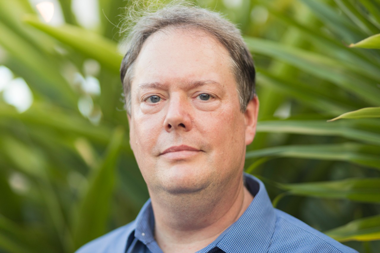A color photograph shwoing a man in a blue shirt with green leaves in the background.