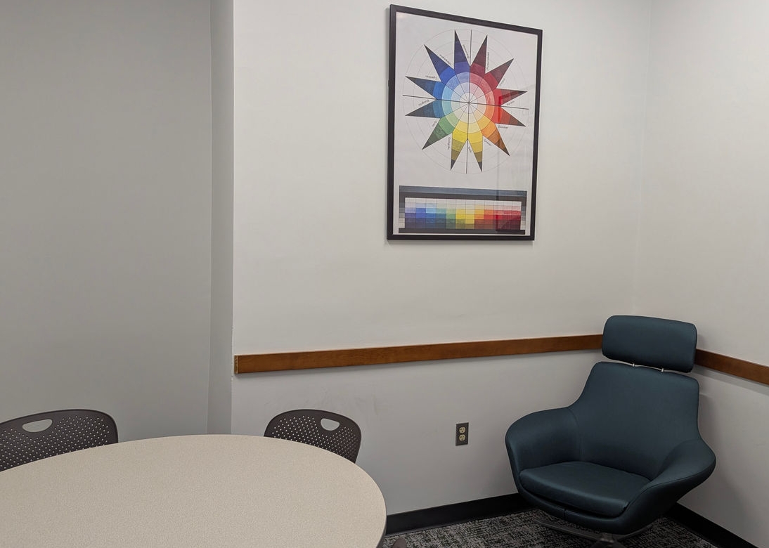 CECH Library study room. In view are two pieces of wall art, a teal armchair, a white round table, and black chairs around the table.
